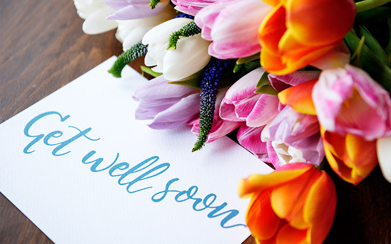 A colorful bouquet of tulips in pink, white, and orange resting on a wooden surface beside a white card with the handwritten message “Get well soon” in blue ink.