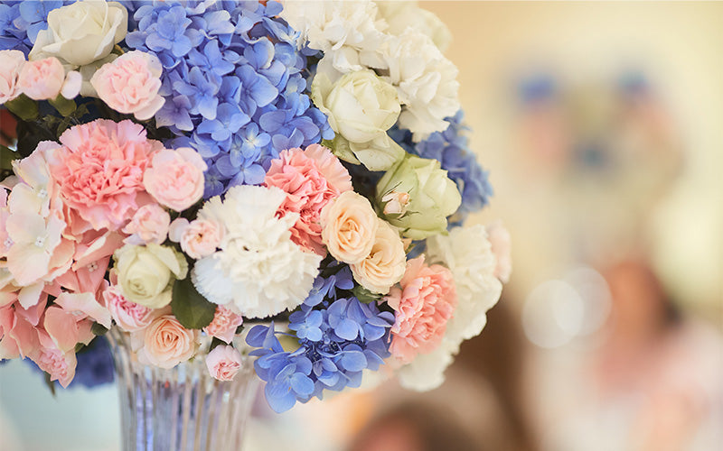 Close-up of floral bouquet with blue hydrangeas, white and peach roses, and pink carnations in glass vase