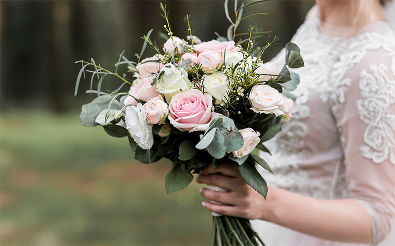 Person in white lace dress holding a bouquet of pink and white roses, ranunculus, and eucalyptus, outdoors.